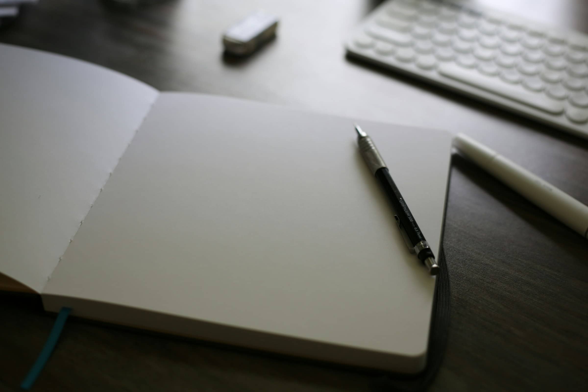 An open blank notebook with a pencil resting on it, keyboard beyond