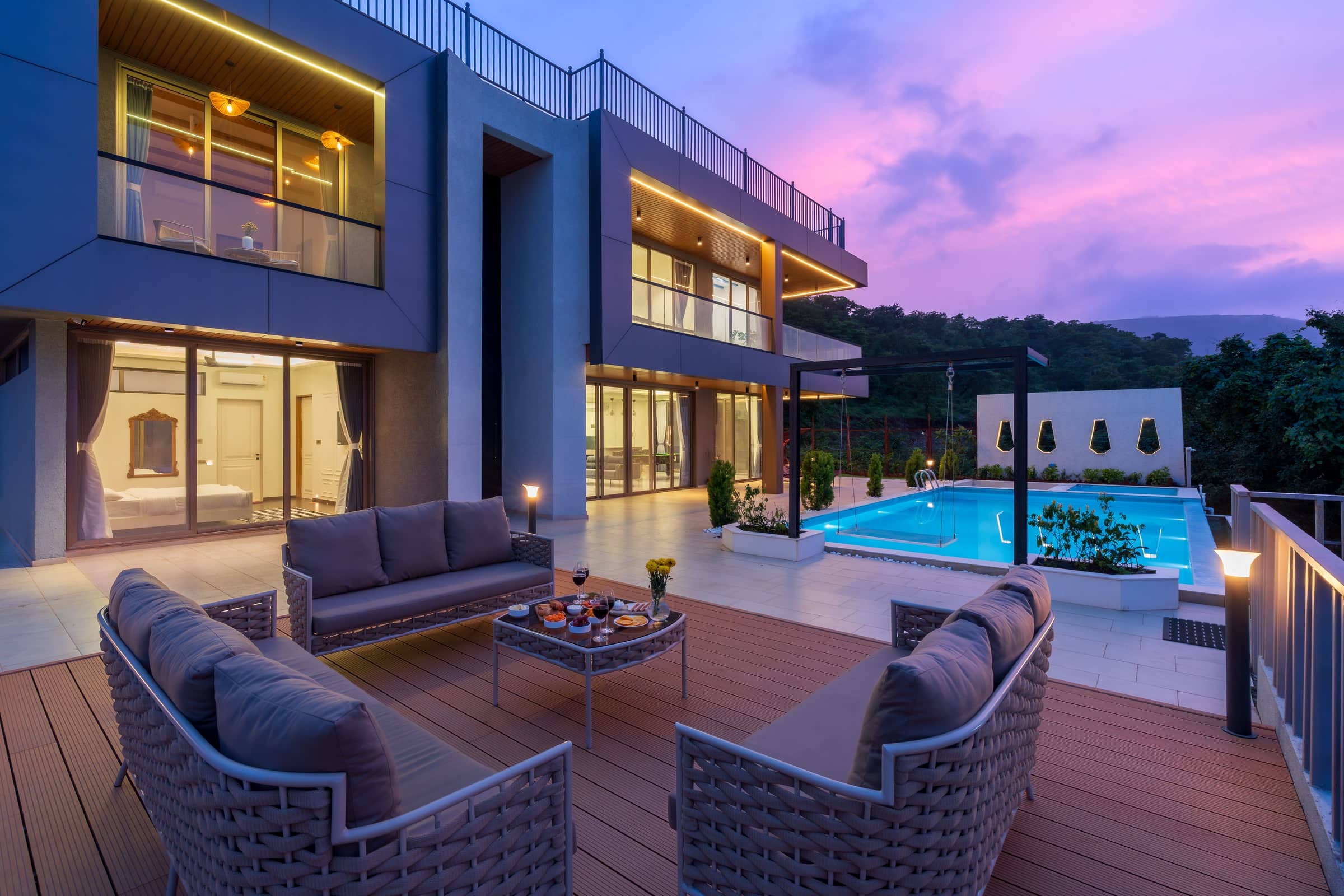 A lit terrace at dusk, seating set out on the deck with the pool and the house glowing behind it