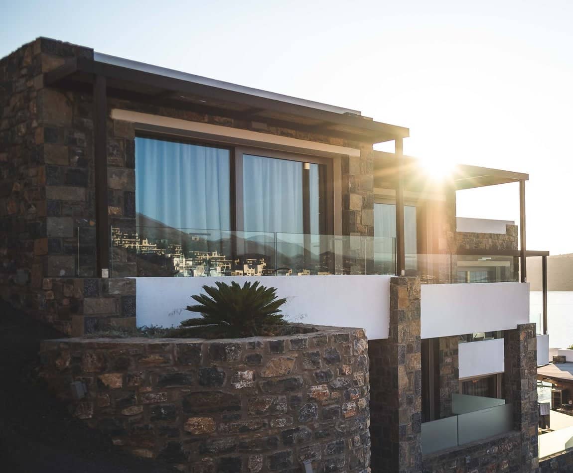 A stone and glass residence on a headland at low sun, its terraces stepping down toward a sheltered bay