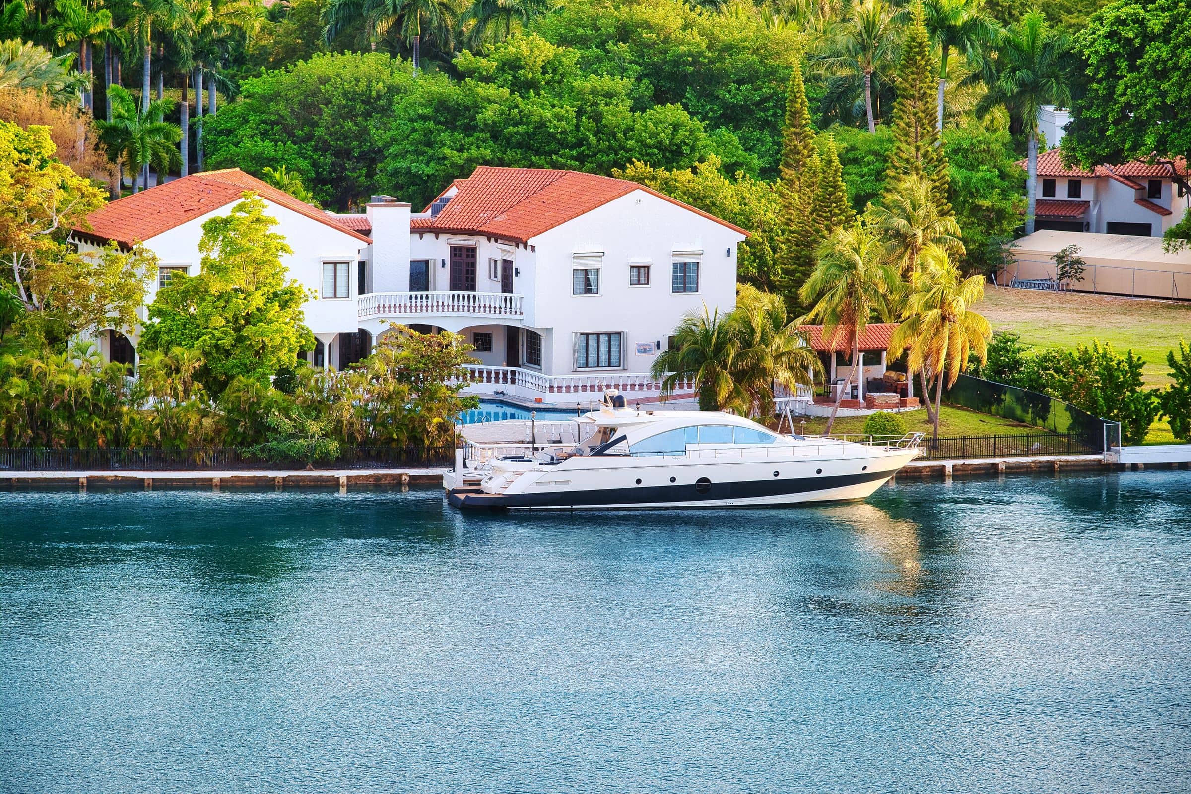 A white waterfront estate with a red tile roof and balconies, a yacht moored at its private dock on calm blue water