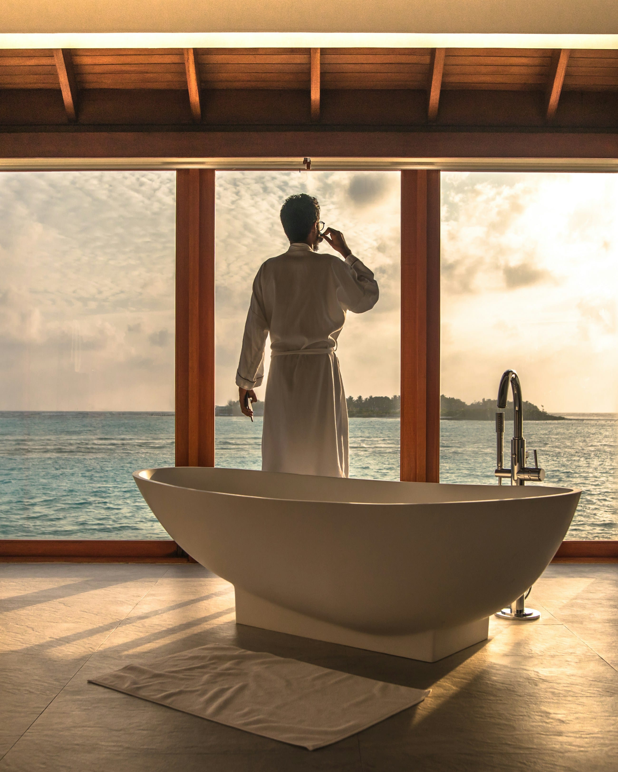 A sculpted freestanding tub set at an open oceanfront window at sunrise, a guest in a white robe looking out across the water