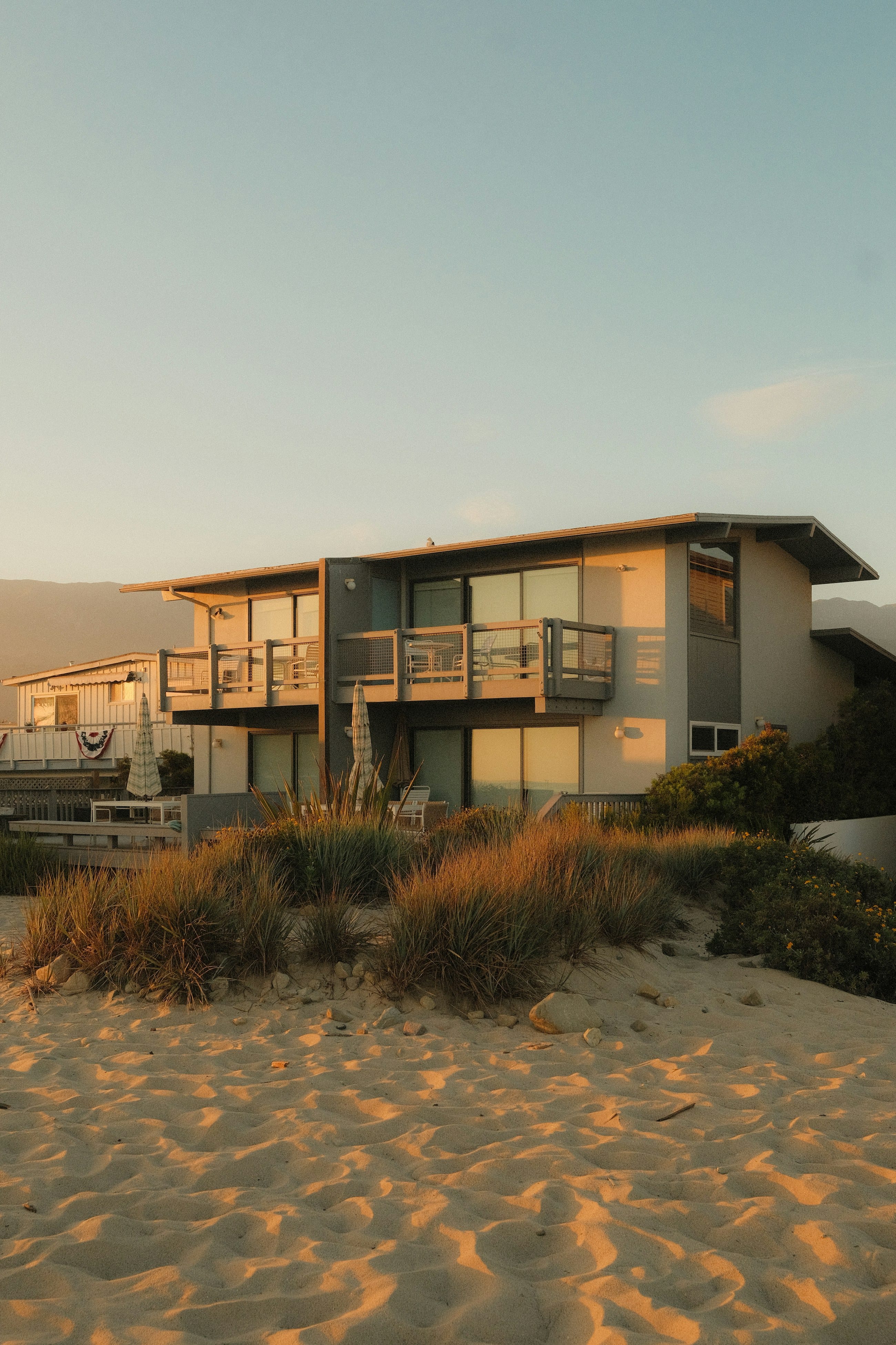 A mid-century beach house standing on a low dune at sunset, its balconies open to the water and sea grass running down to the sand