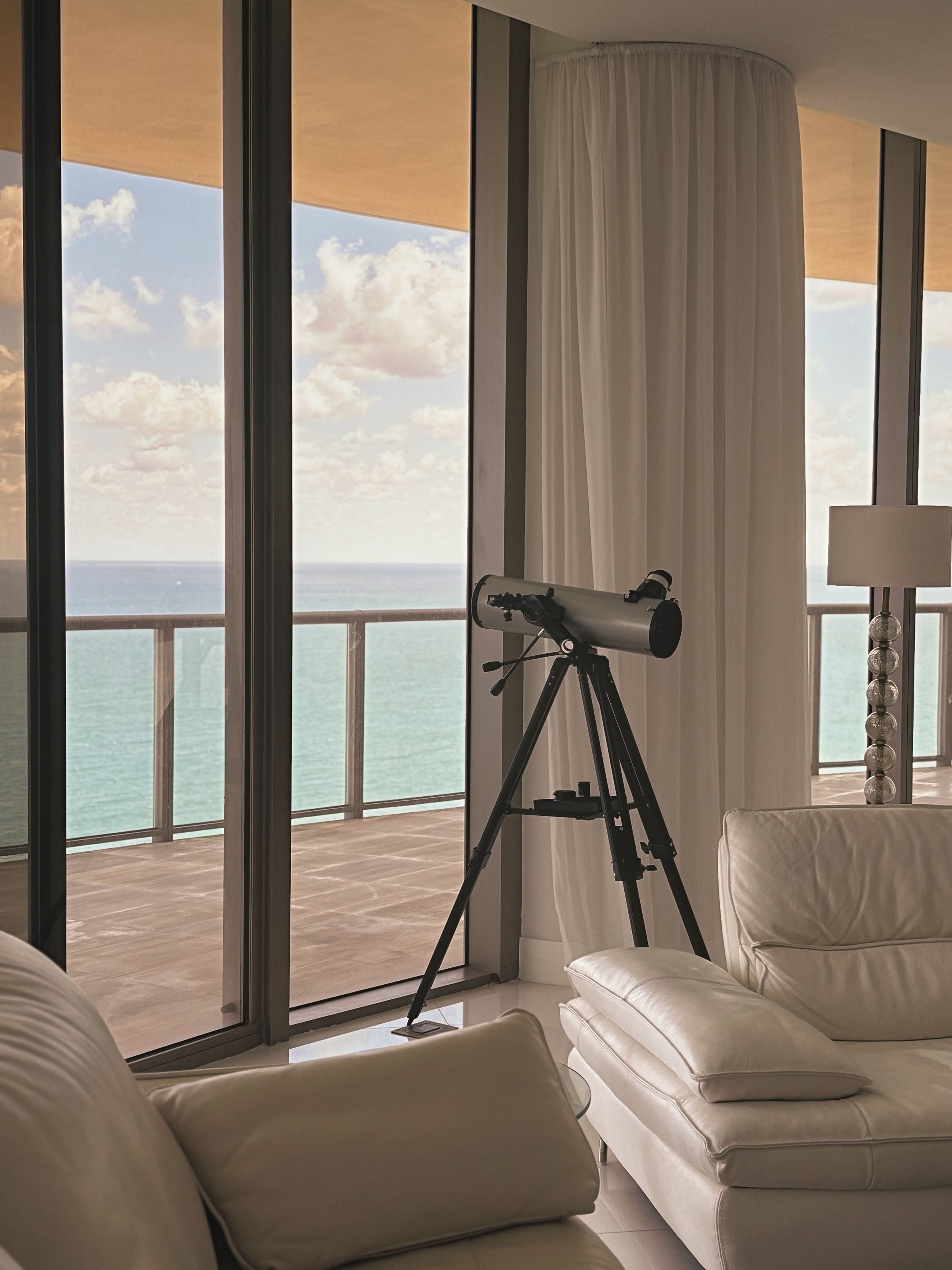 A coastal living room behind full-height glass at golden hour, pale seating turned toward a terrace and the open ocean