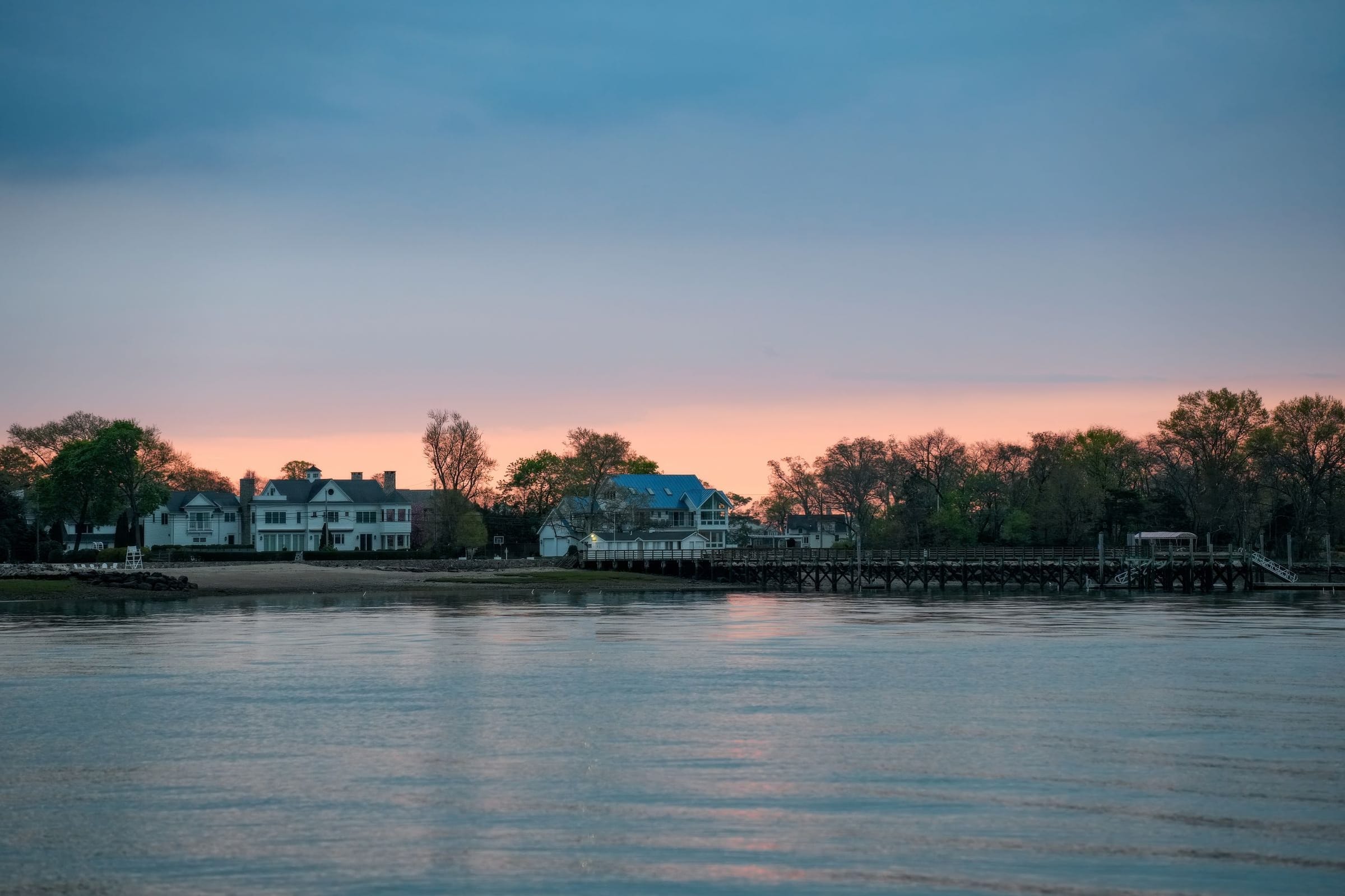 White waterfront houses behind a long wooden dock on still water at dusk, an afterglow over the treeline