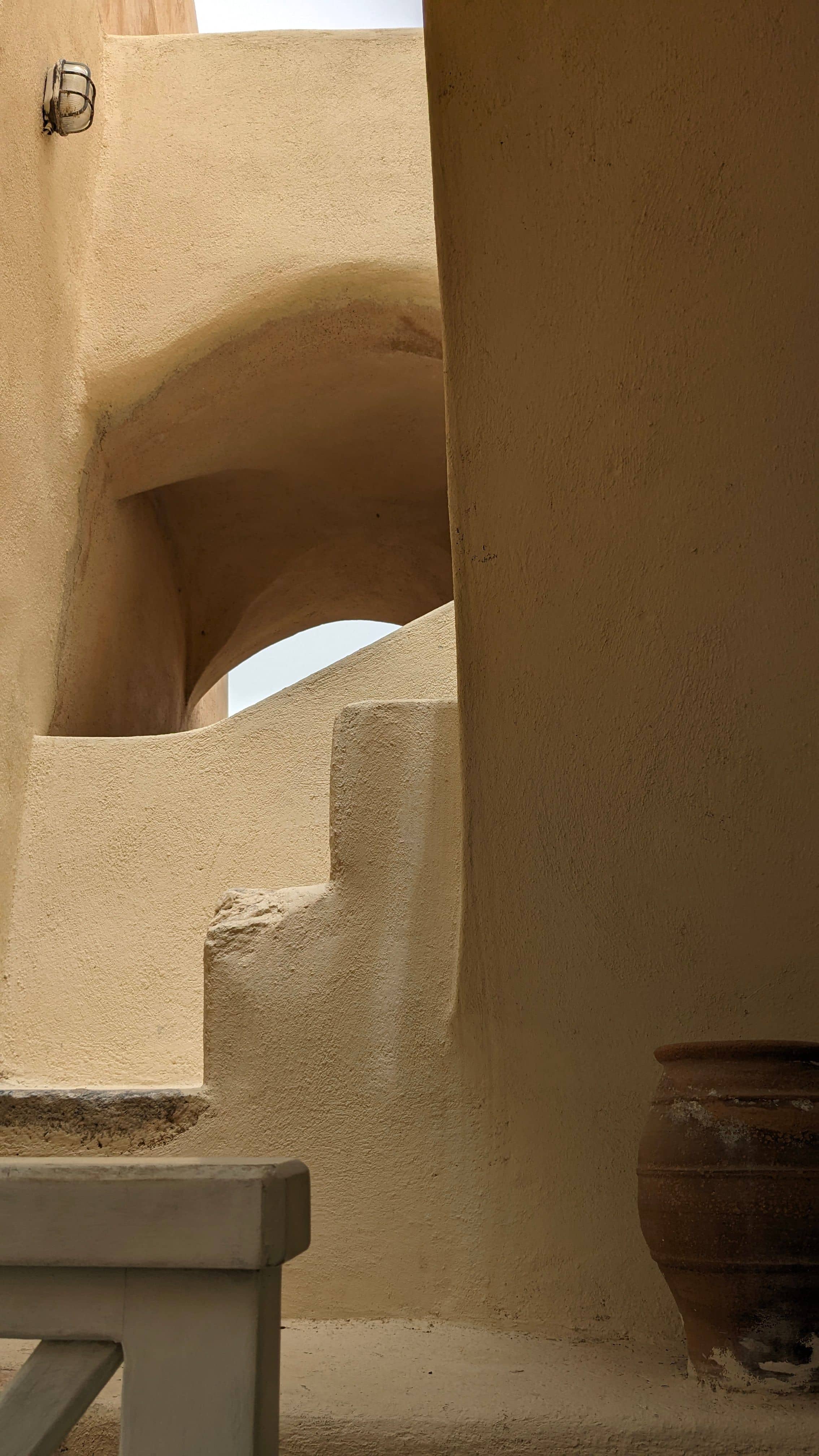 Sunlit sand-plaster archways and steps with a terracotta urn, warm and quiet