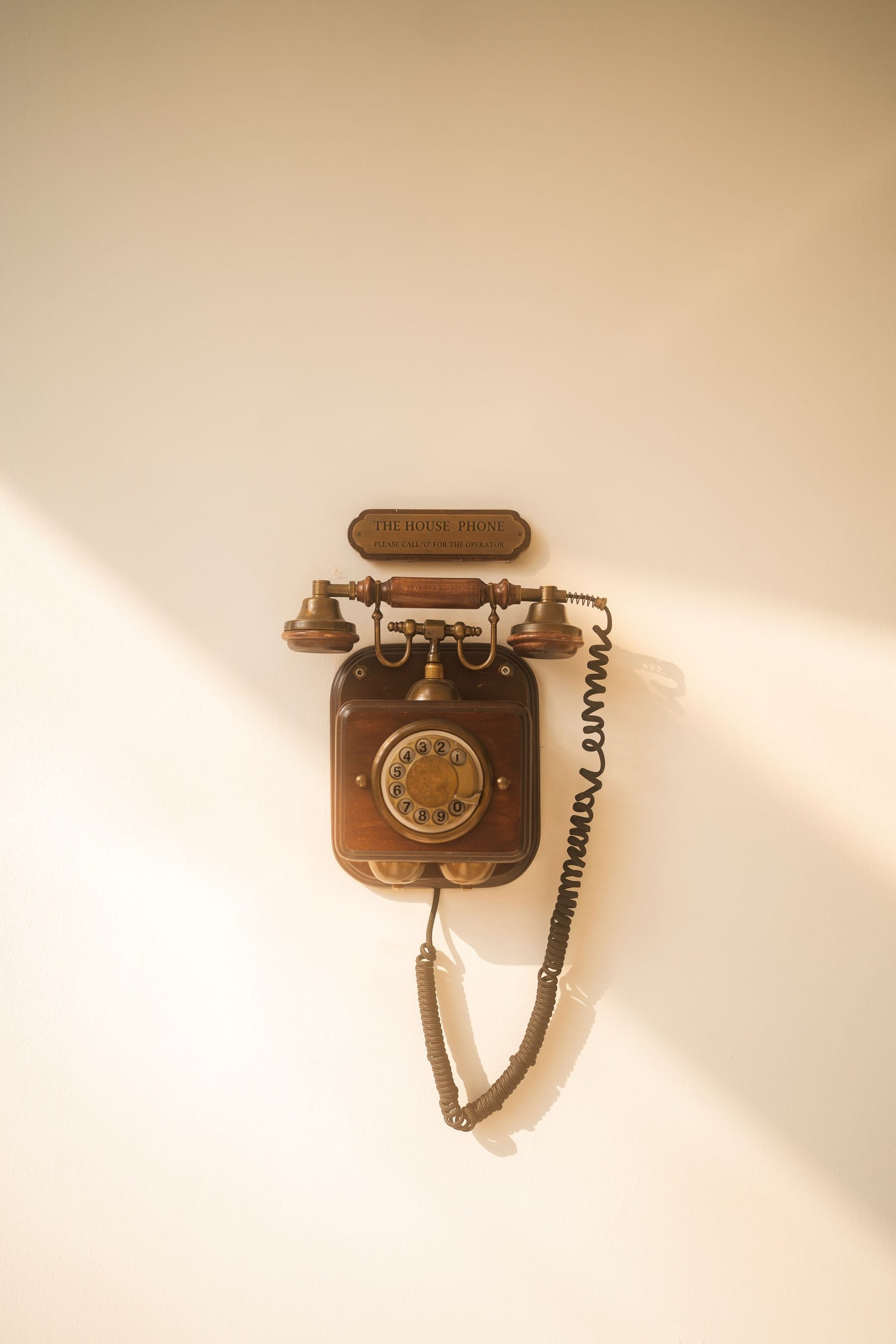 A vintage wall-mounted house telephone in wood and brass on a warm cream wall, soft daylight falling across it
