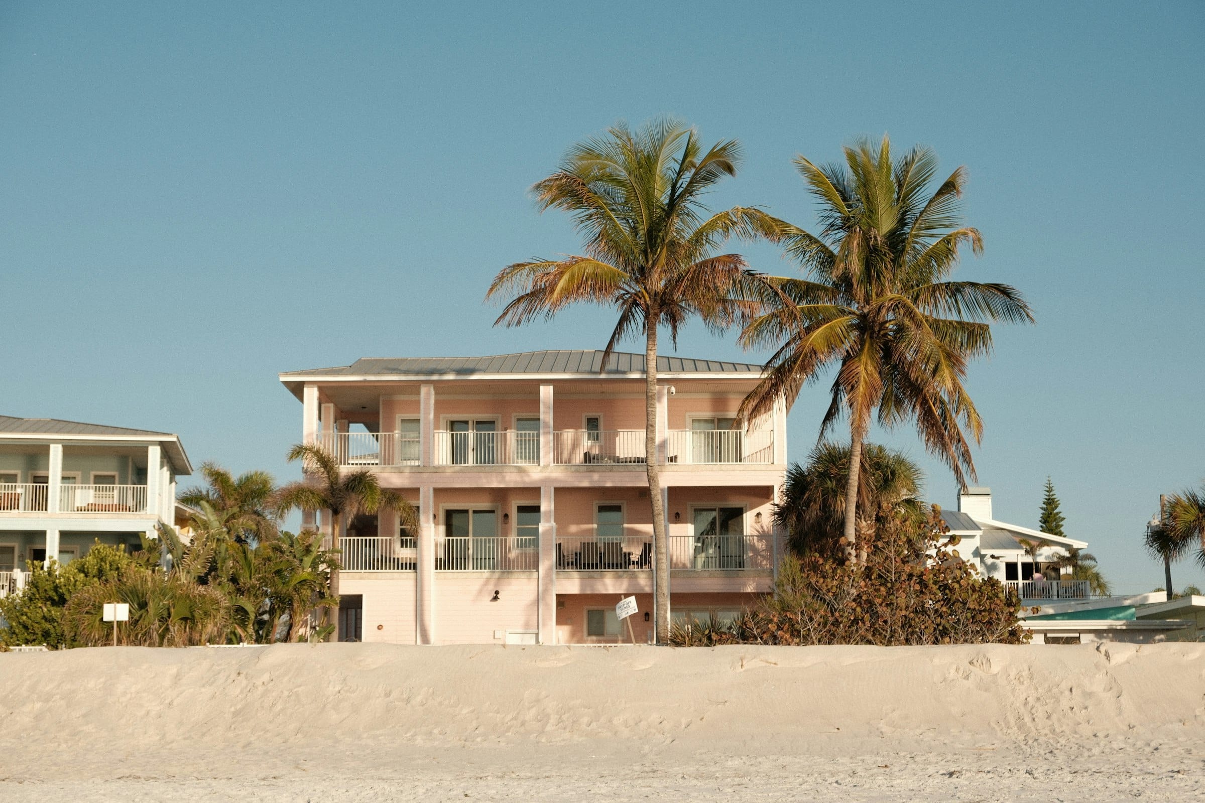 Pink three-story beach house with white railings behind the dune, palms overhead