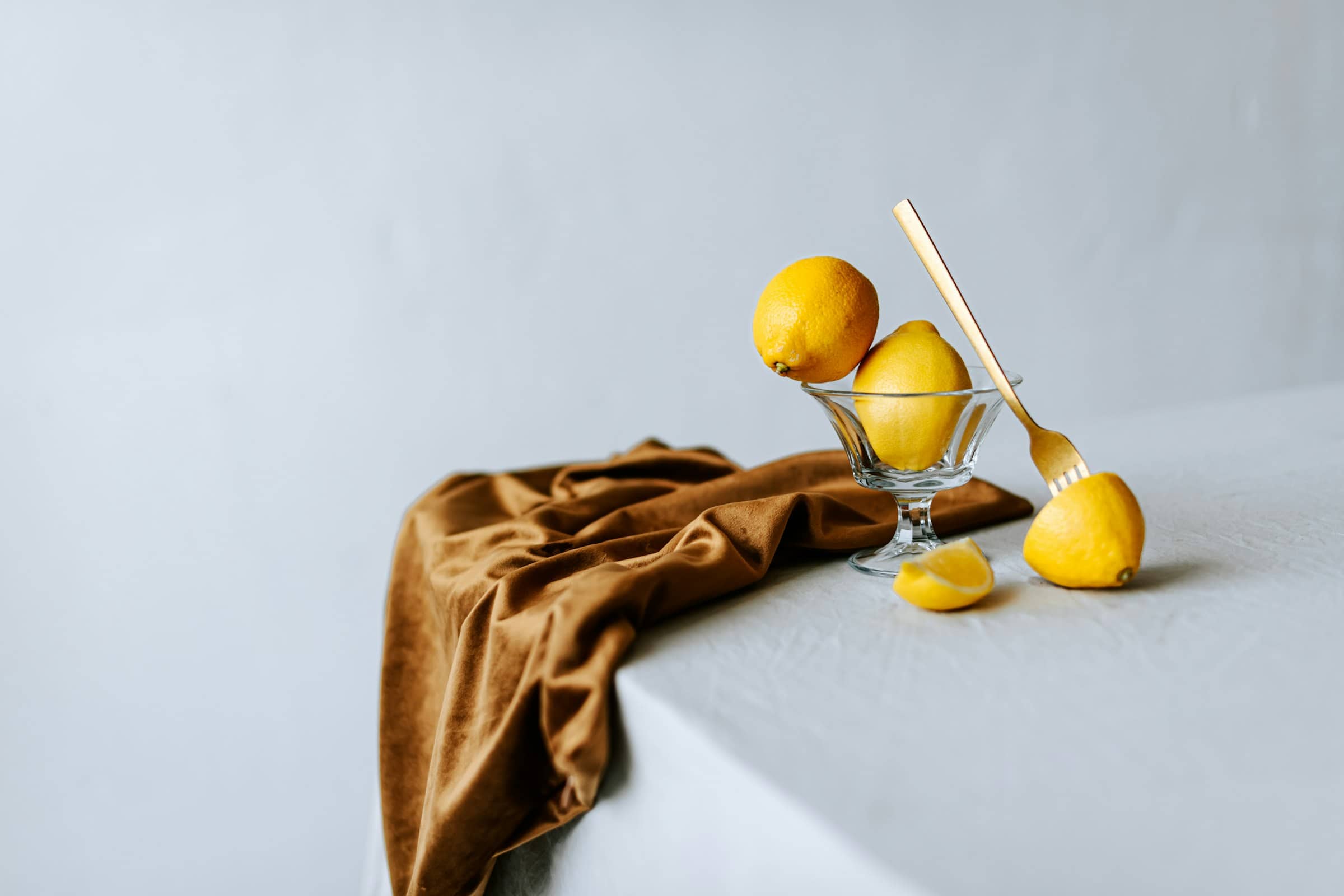 Lemons in a glass pedestal bowl on pale linen, an ochre cloth alongside