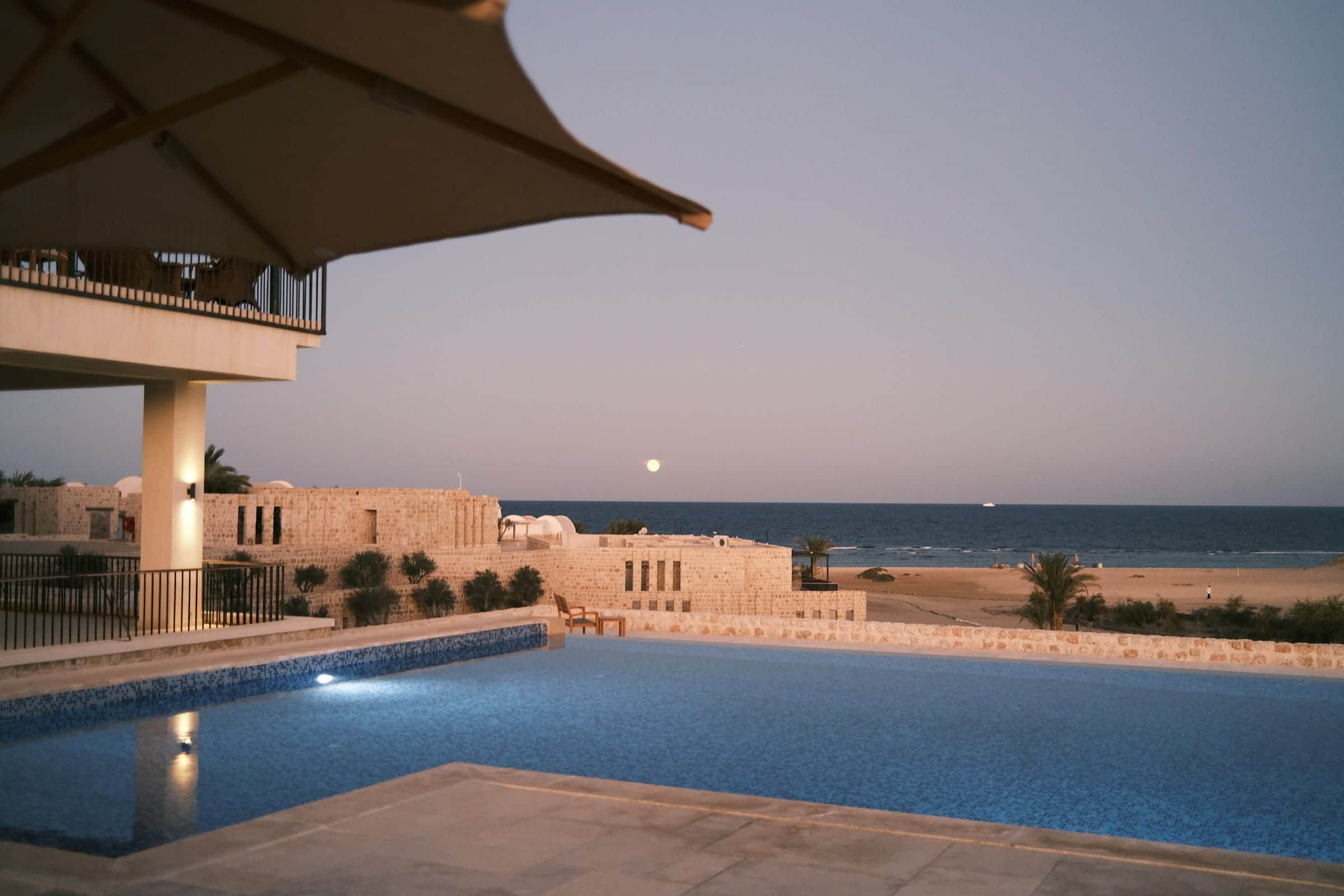 A tiled pool at dusk before low buildings of pale stone, a full moon rising over the calm sea beyond the sand