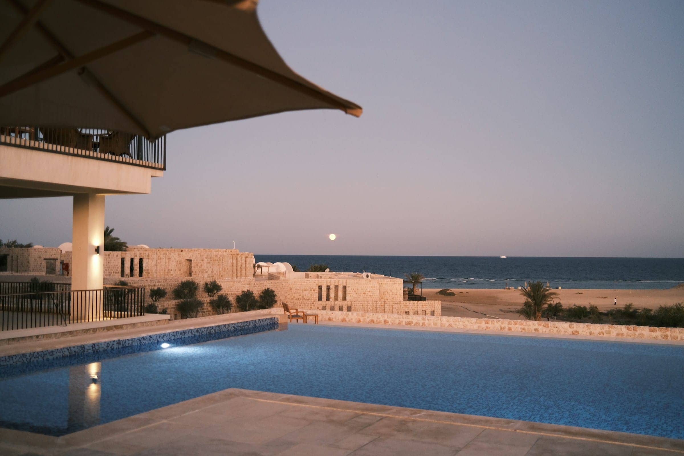 A tiled pool at dusk before low buildings of pale stone, a full moon rising over the calm sea beyond the sand