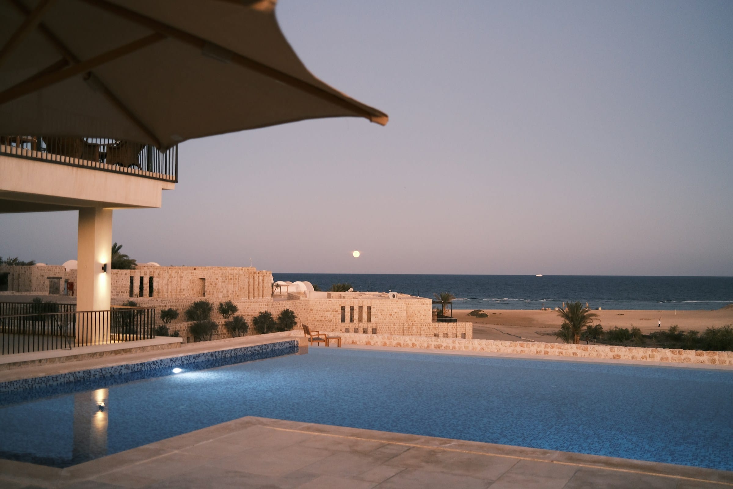 A tiled pool at dusk before low buildings of pale stone, a full moon rising over the calm sea beyond the sand