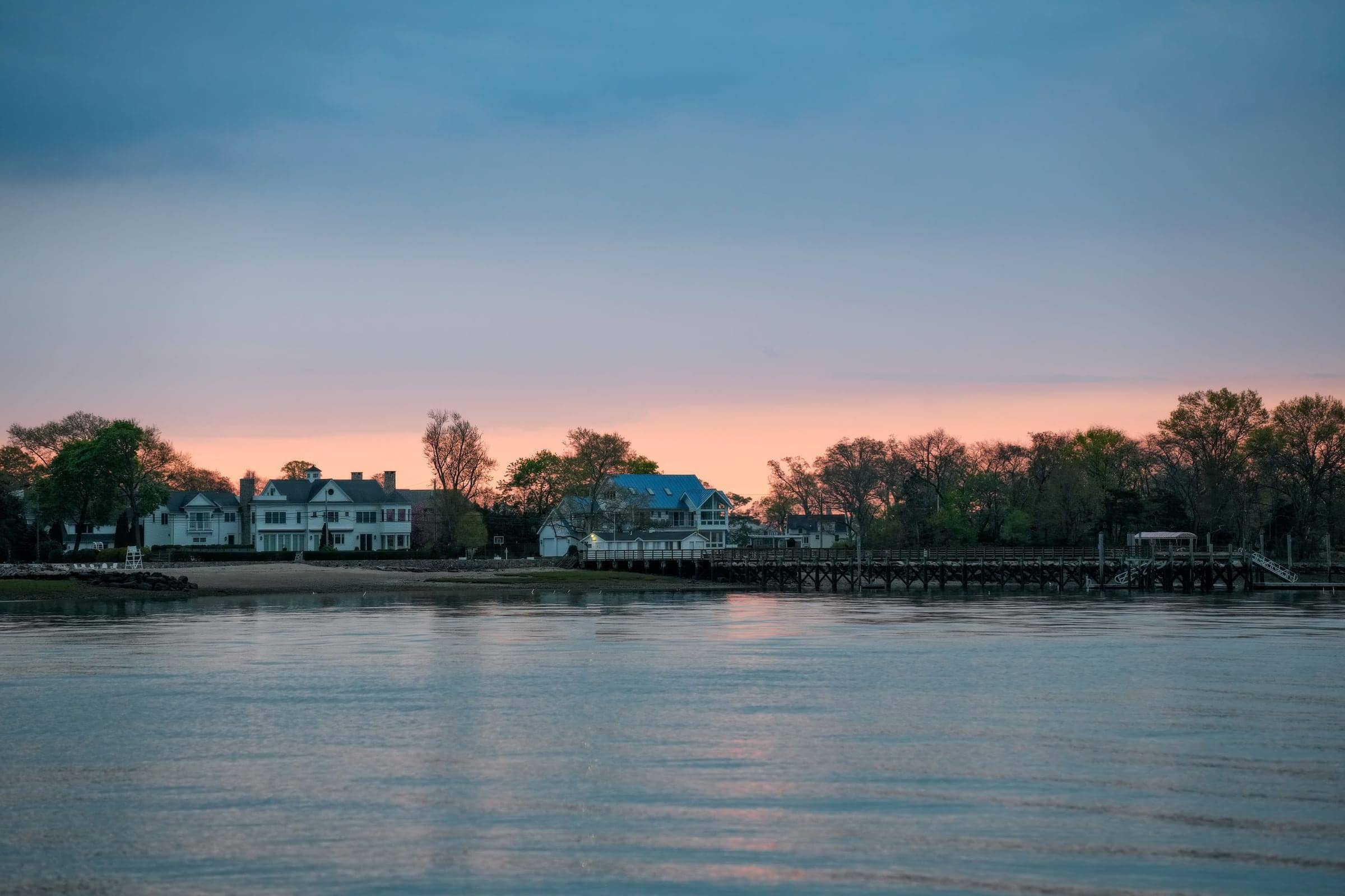 White waterfront houses behind a long wooden dock on still water at dusk, an afterglow over the treeline