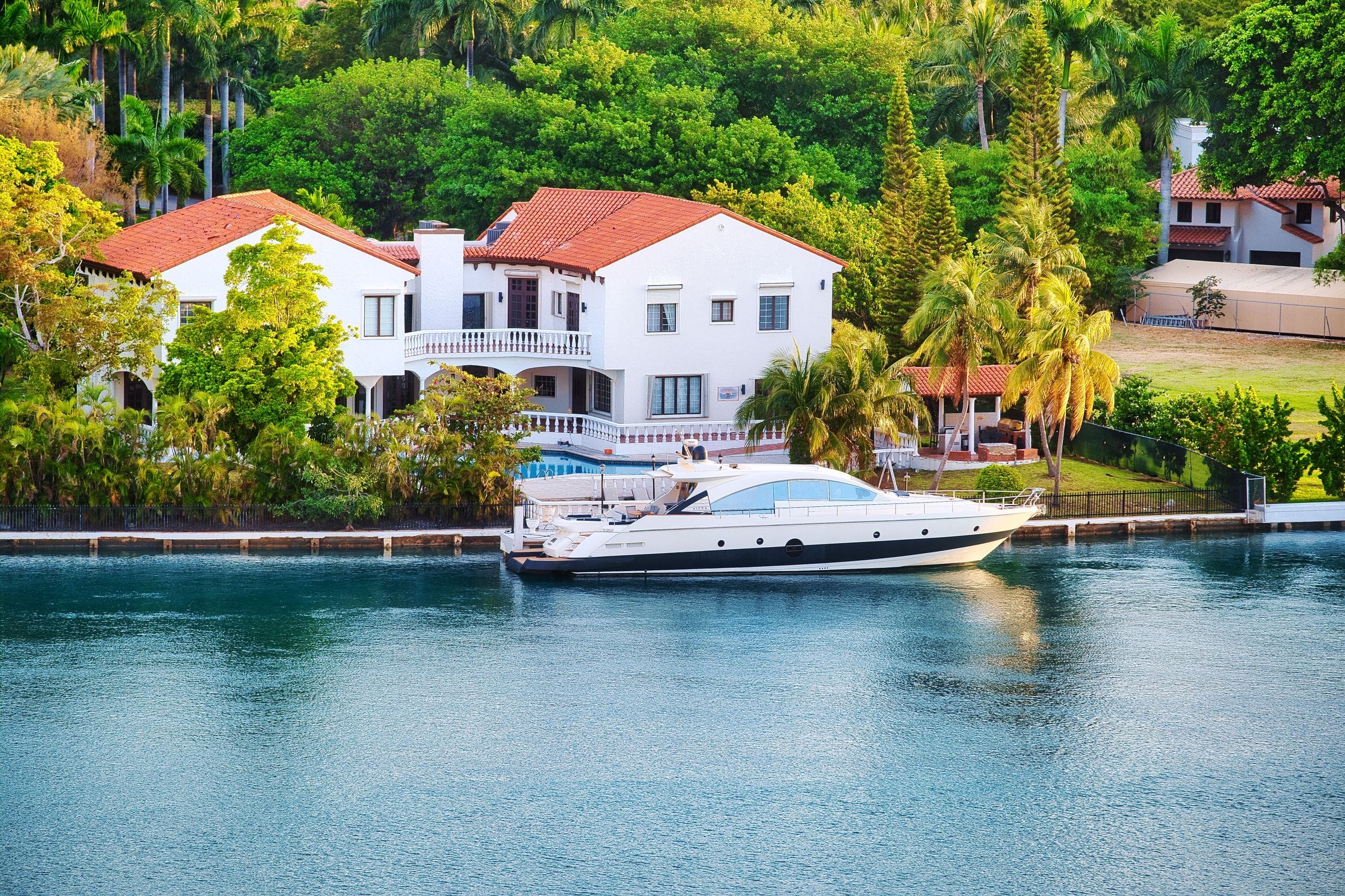 A white waterfront estate with a red tile roof and balconies, a yacht moored at its private dock on calm blue water